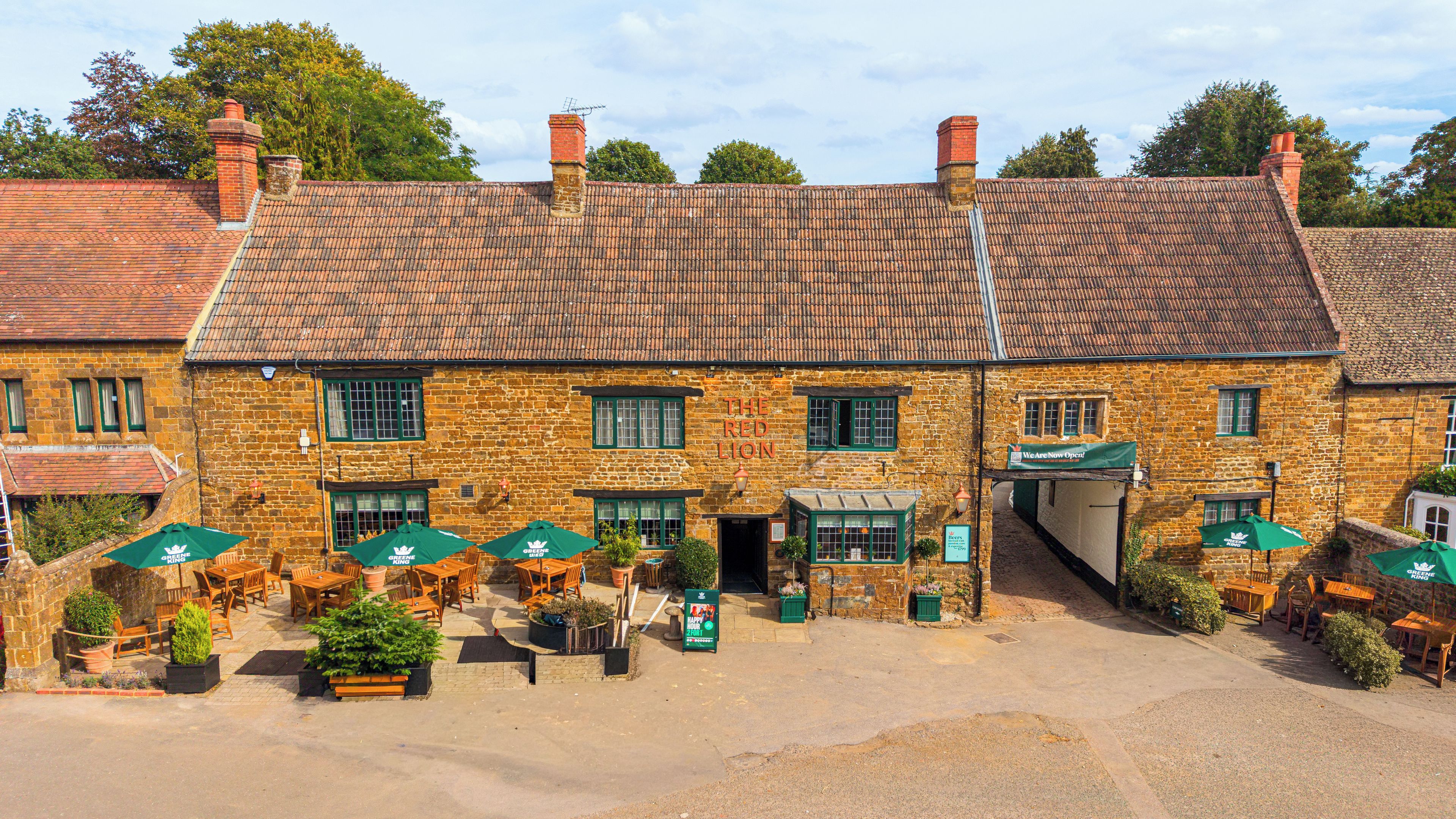 A traditional stone building with a tiled roof, outdoor seating with green umbrellas, and a sign indicating it is open.