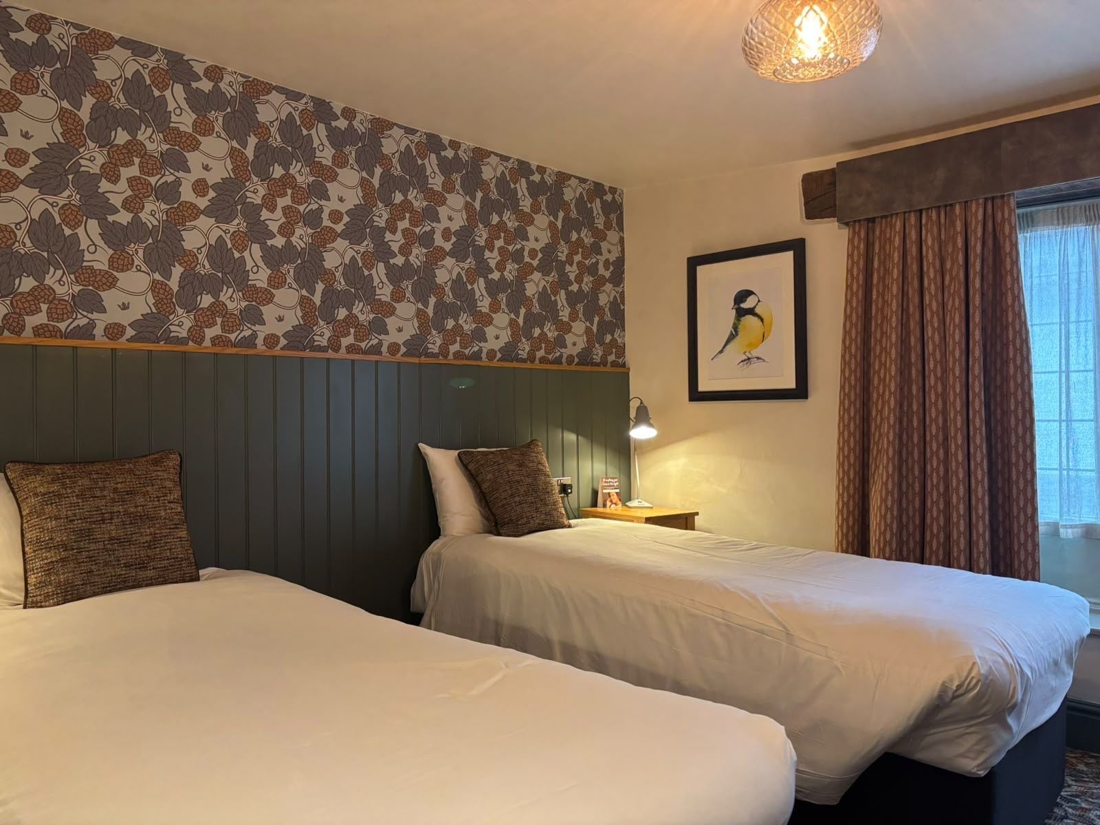 Ein Hotelzimmer mit zwei Betten, einer Wandlampe, einem Bilderrahmen mit einem Vogel und einem Fenster mit Vorhängen.