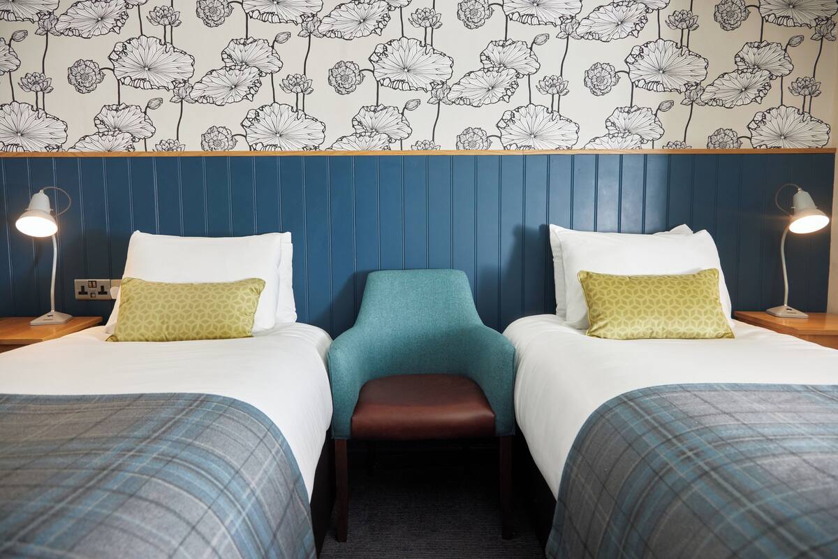 Habitació d'hotel amb dos llits, una cadira color turquesa i un fons de pantalla amb motius florals.
