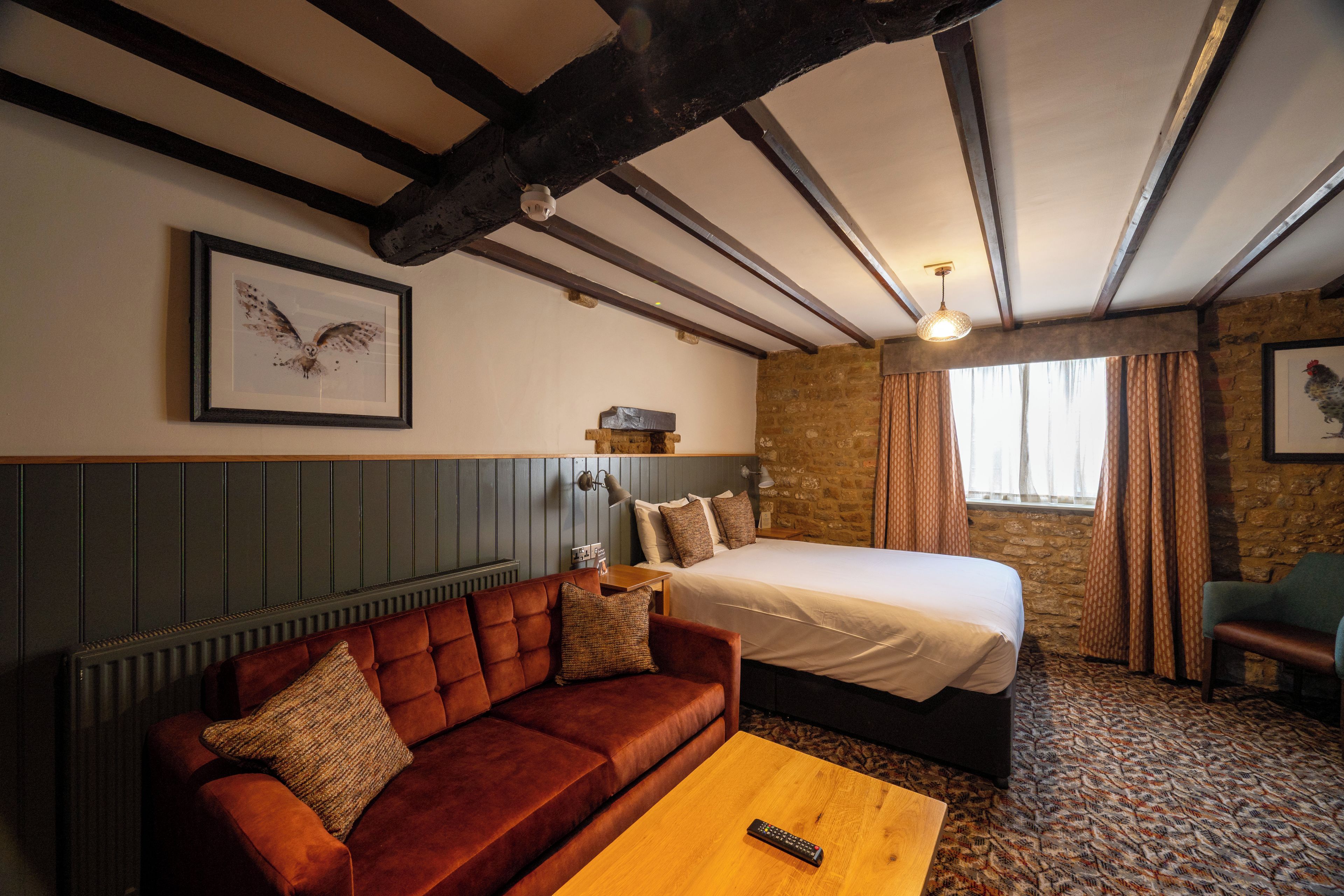 Quarto de hotel com cama, sofá, mesa de centro de madeira, luminária fixada na parede e quadro com a imagem de uma coruja.