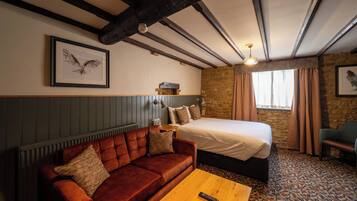 Quarto de hotel com cama, sofá, mesa de centro de madeira, luminária fixada na parede e quadro com a imagem de uma coruja.
