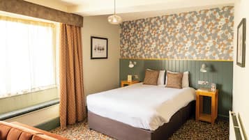 A hotel room with a bed, a bedside table, a nightstand, a window with curtains, a wall with a leaf pattern, and a framed picture on the wall.