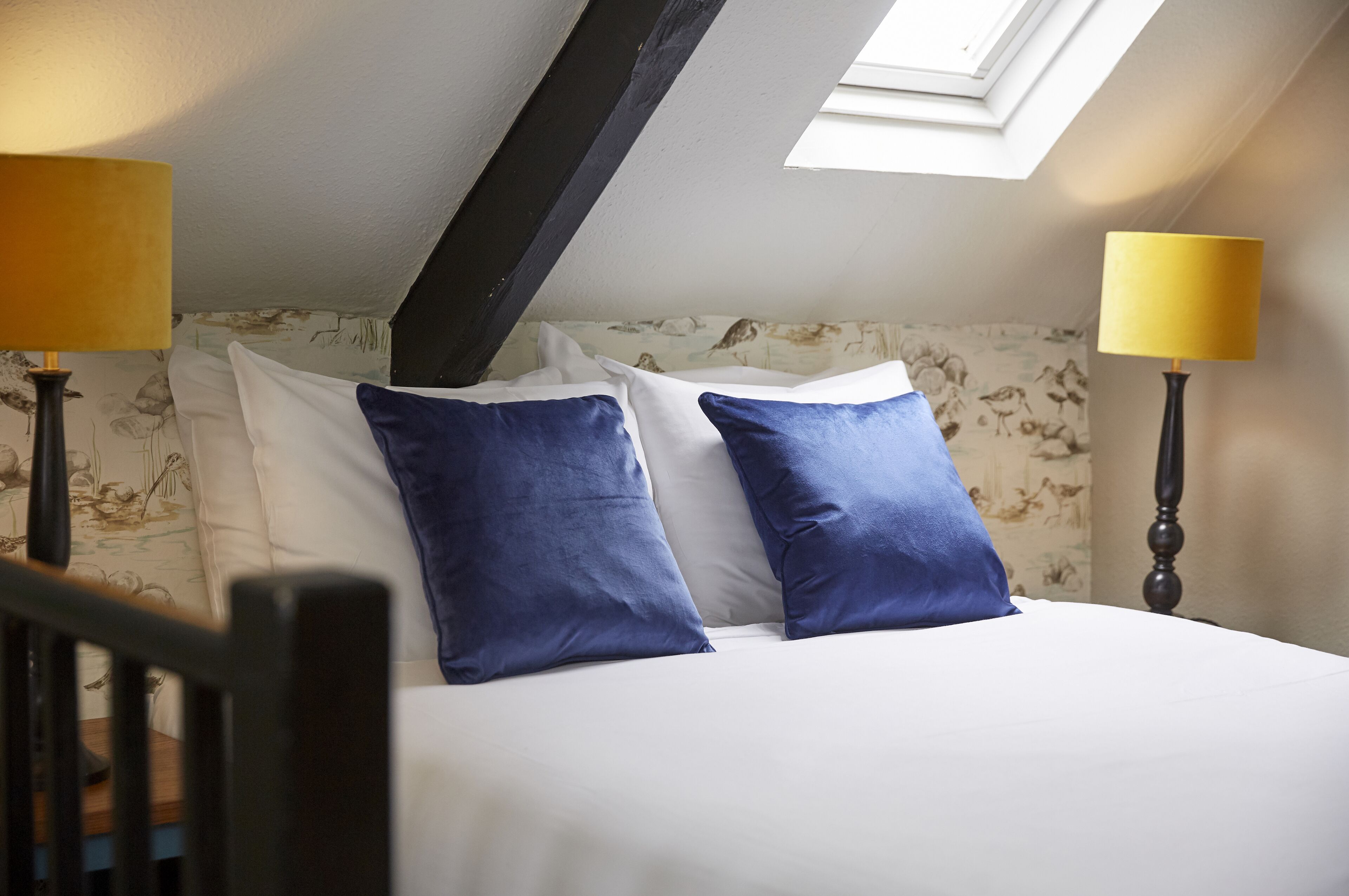 A neatly made bed with white and blue pillows, two lamps on either side, and a skylight above.