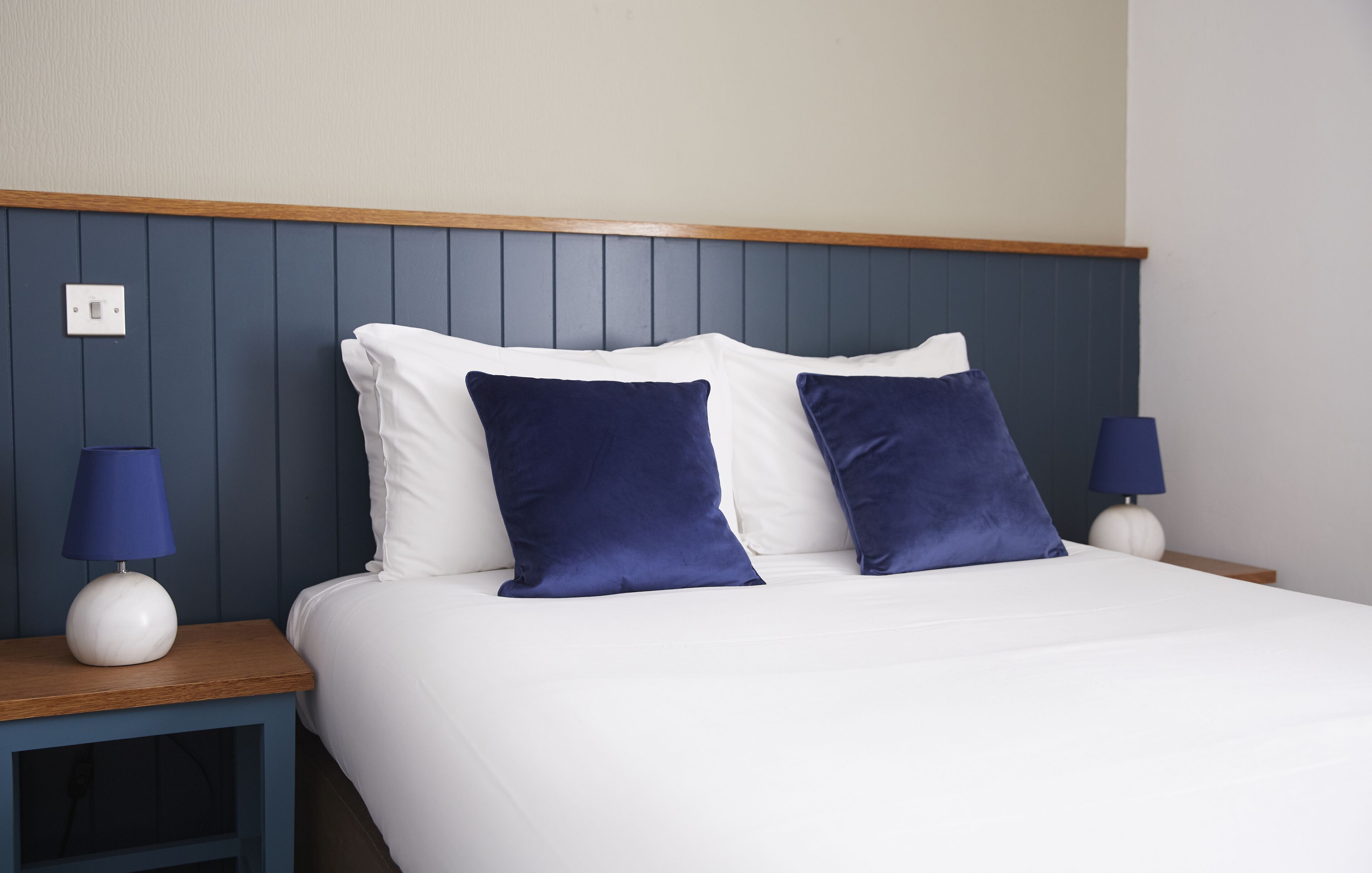 A neatly made bed with white and dark blue pillows, two bedside lamps, and a wooden headboard.