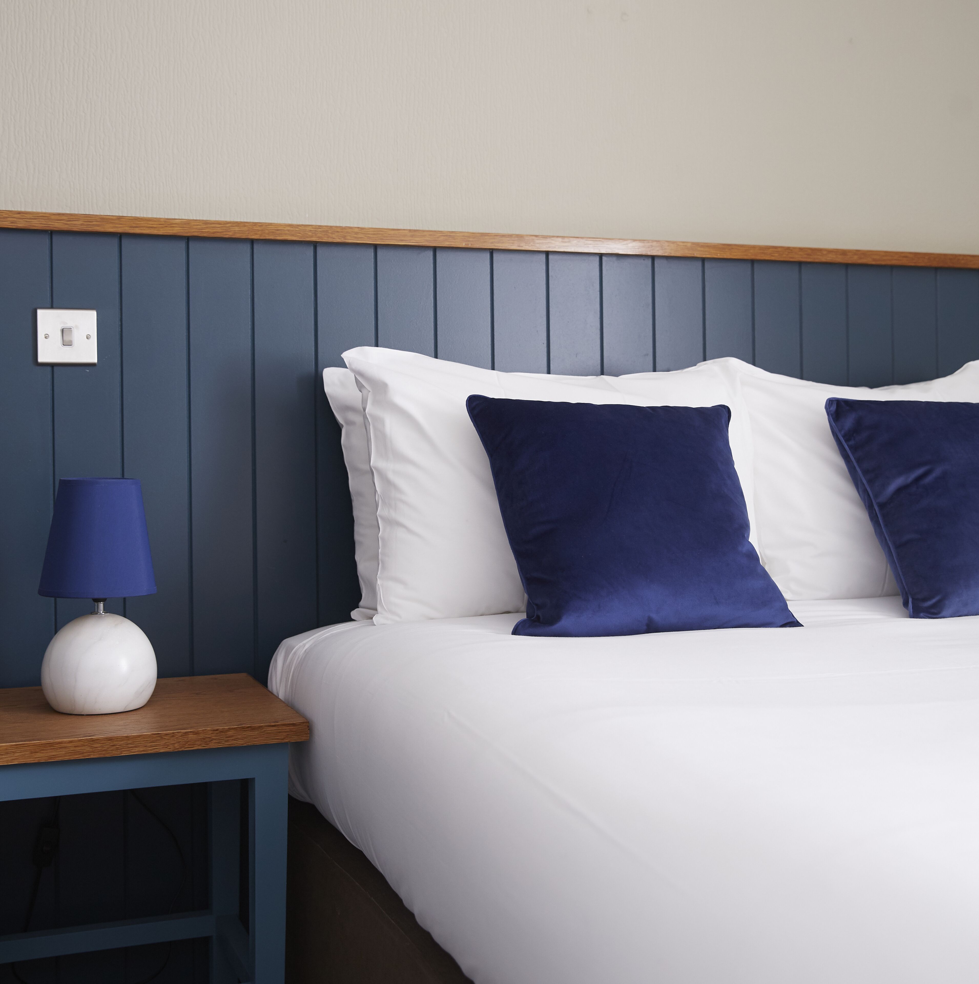 A neatly made bed with white linens and two dark blue pillows, a wooden nightstand with a blue lamp, and a wall switch.