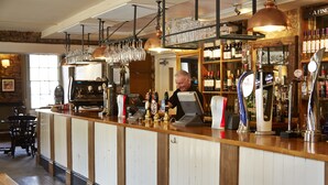 Un espace bar avec un comptoir, des tabourets de bar et un barman. On y trouve des étagères remplies de bouteilles et de verres.