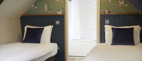 Un dormitorio con dos camas, cada una con ropa de cama blanca y almohadas azul oscuro. La habitación tiene un techo inclinado, decoración de aves en la pared y una zona de ducha visible.