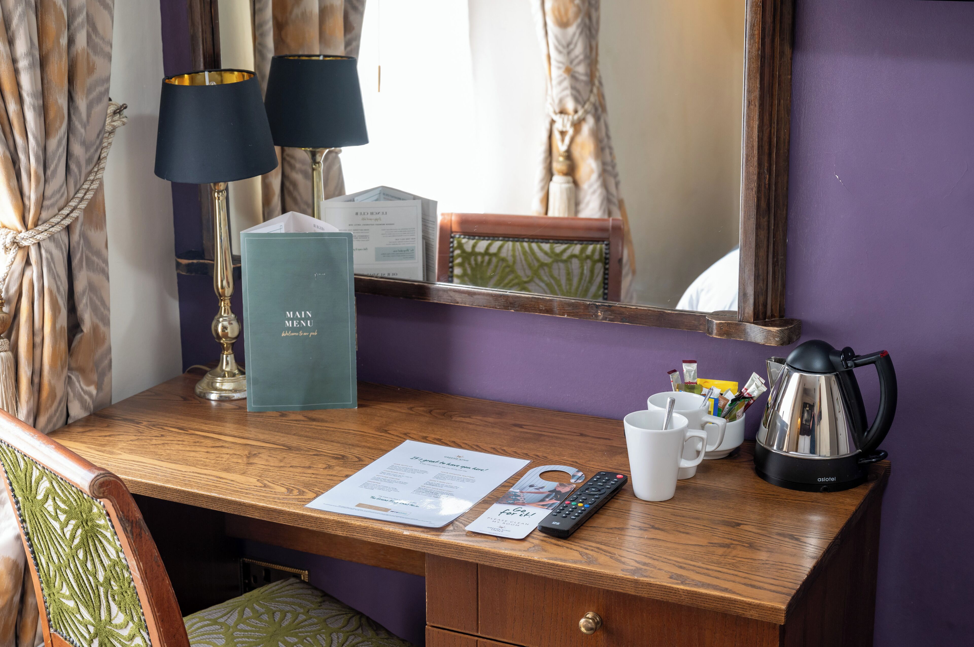 A wooden desk with a remote control, a menu, a kettle, and a cup.