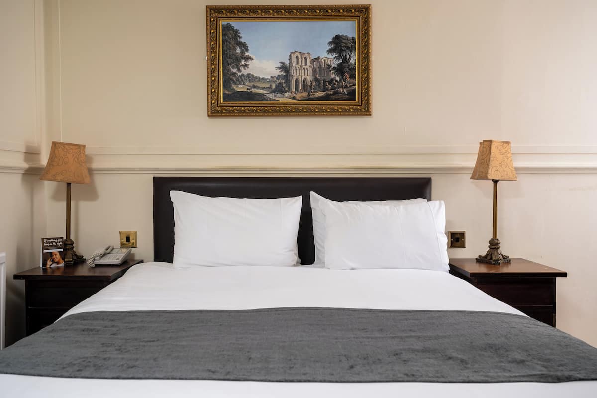 Ein Hotelzimmer mit einem großen Bett, zwei Nachttischlampen, einem gerahmten Bild an der Wand und einem Nachttisch mit Telefon und einer Kleinigkeit.