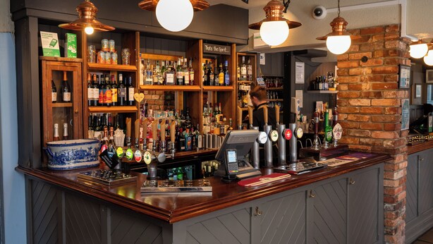 A bar area with wooden shelves stocked with various bottles, a cash register, and a beer tap. - Antelope Hotel by Greene King Inns (Poole)