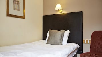 En snyggt bäddad säng med vita lakan och en enda grå kudde, en väggmonterad lampa och en röd, mönstrad stol i ett hotellrum.