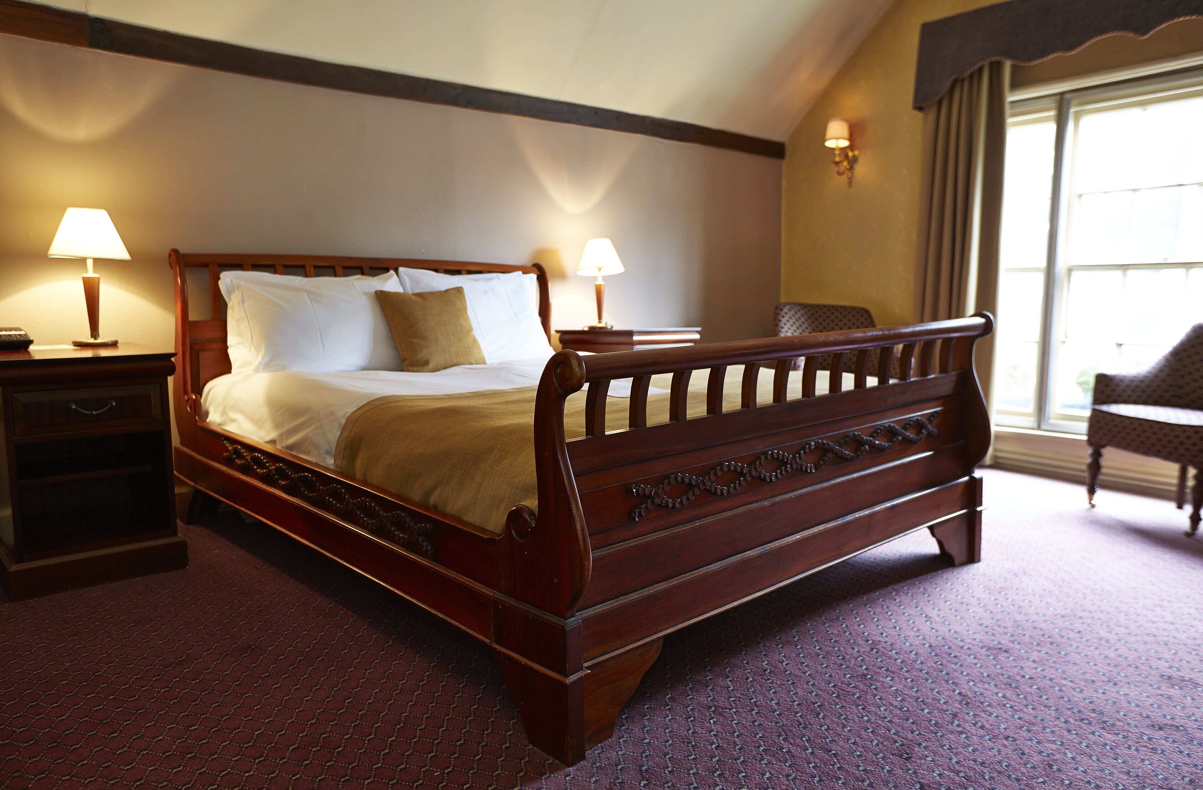 Quarto de hotel com cama de madeira, duas luminárias de cabeceira, mesa de apoio, uma cadeira e janela com cortinas.