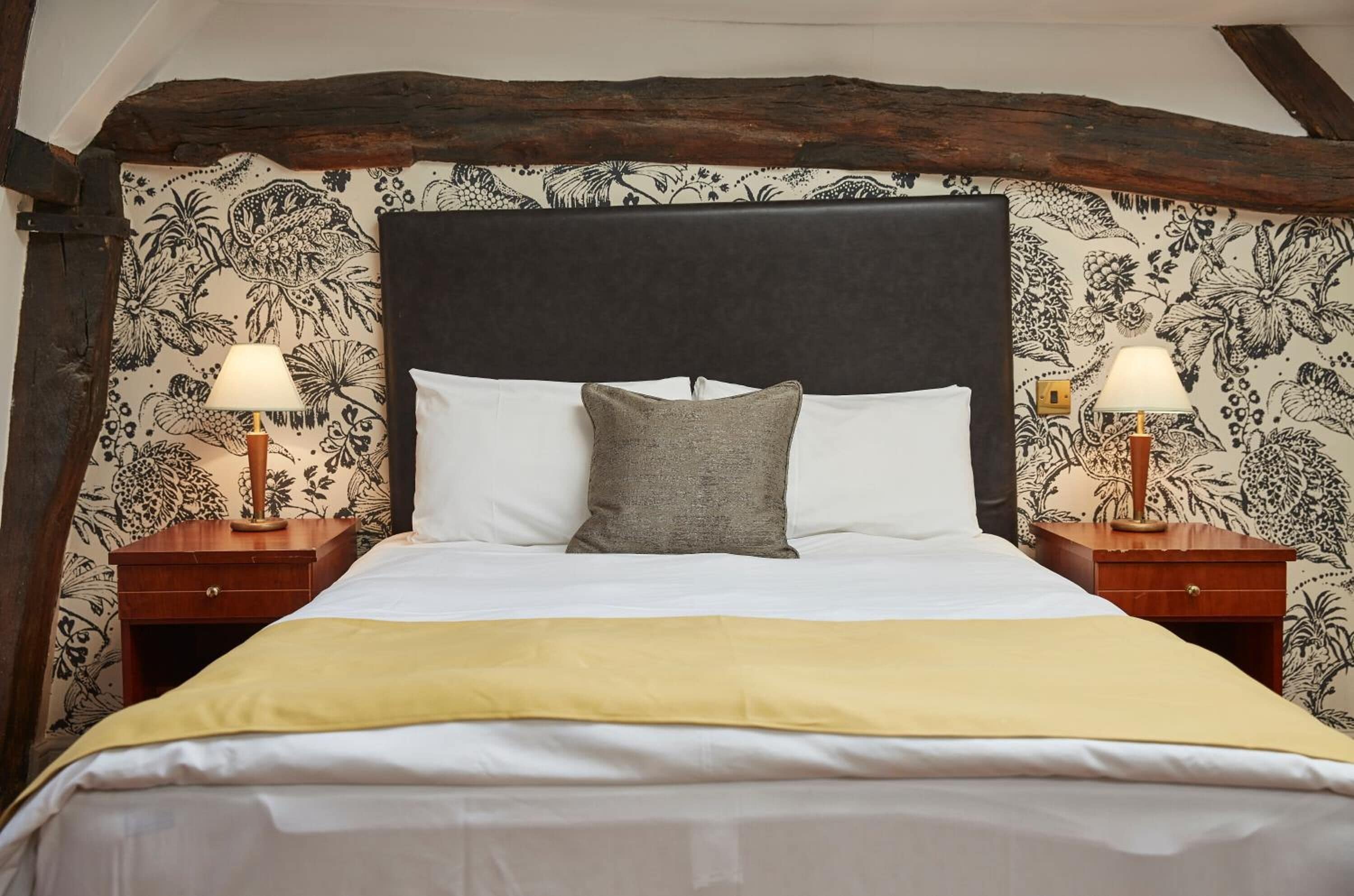 A bed with a dark headboard, white bedding, and a yellow throw blanket. There are two wooden nightstands with lamps on either side of the bed.