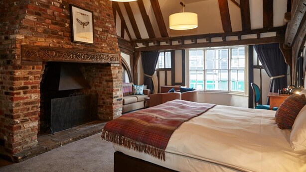 A traditional room with exposed brick walls, a fireplace, a bed with a plaid blanket, and large windows with curtains. - White Hart Hotel by Greene King Inns (Colchester)