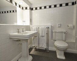 Un baño con lavamanos, espejo y retrete blancos. Hay dos toallas blancas colgadas en un perchero.