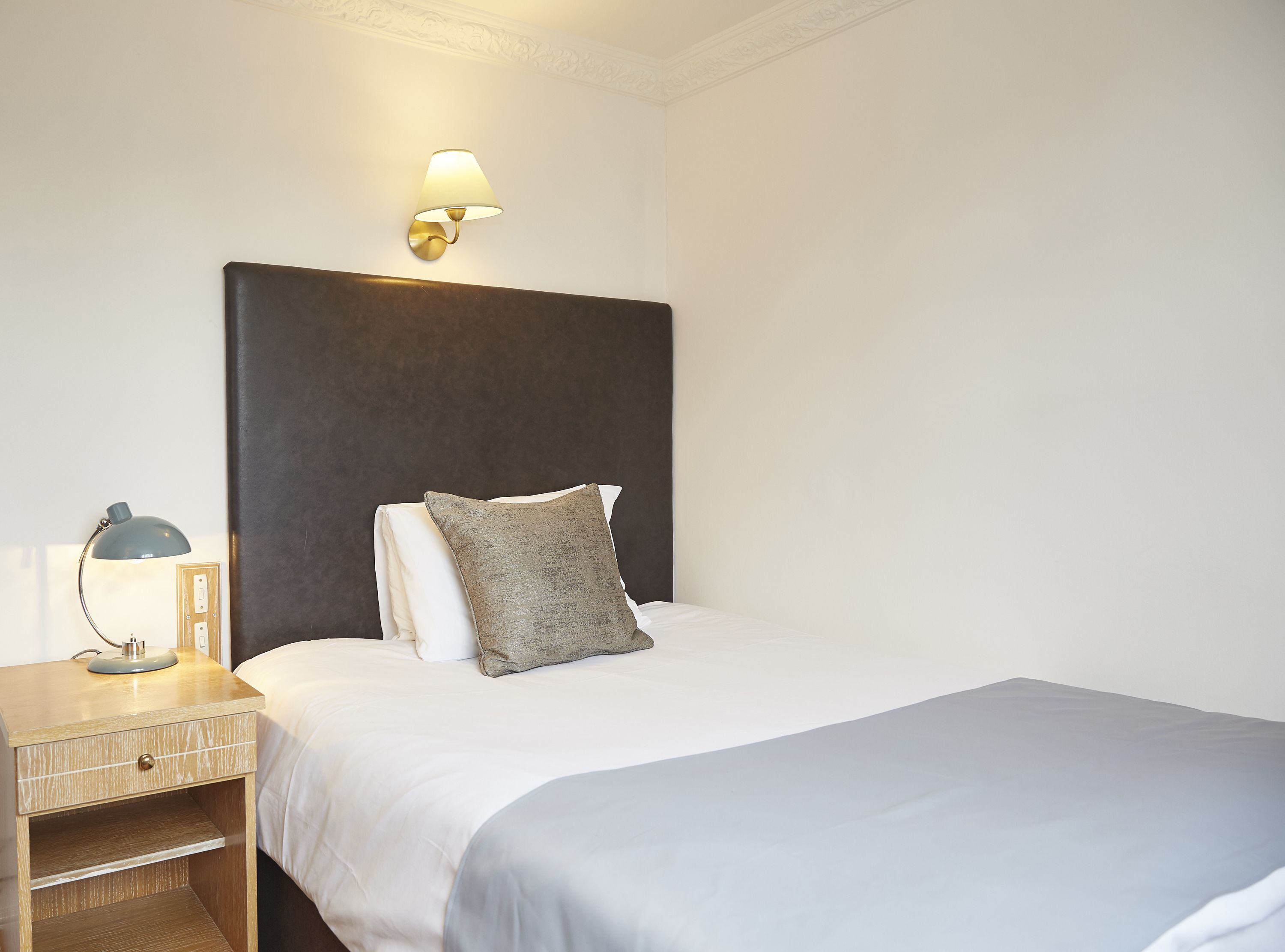 A neatly made bed with a grey pillow and a white duvet, a bedside lamp, and a wall-mounted light fixture.