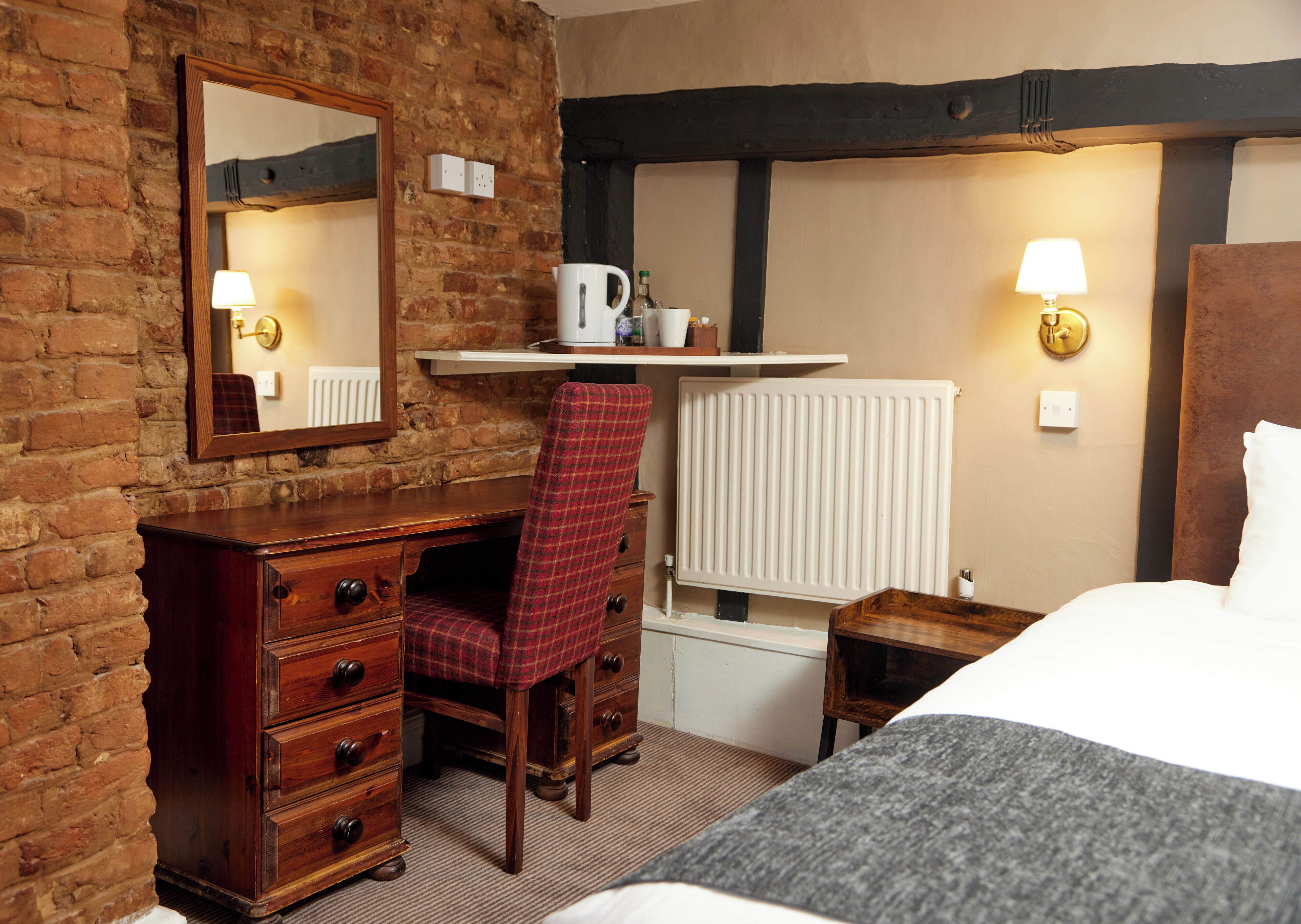 A hotel room with a wooden dresser, a plaid chair, a desk with a kettle, a radiator, and a bed with a grey blanket.