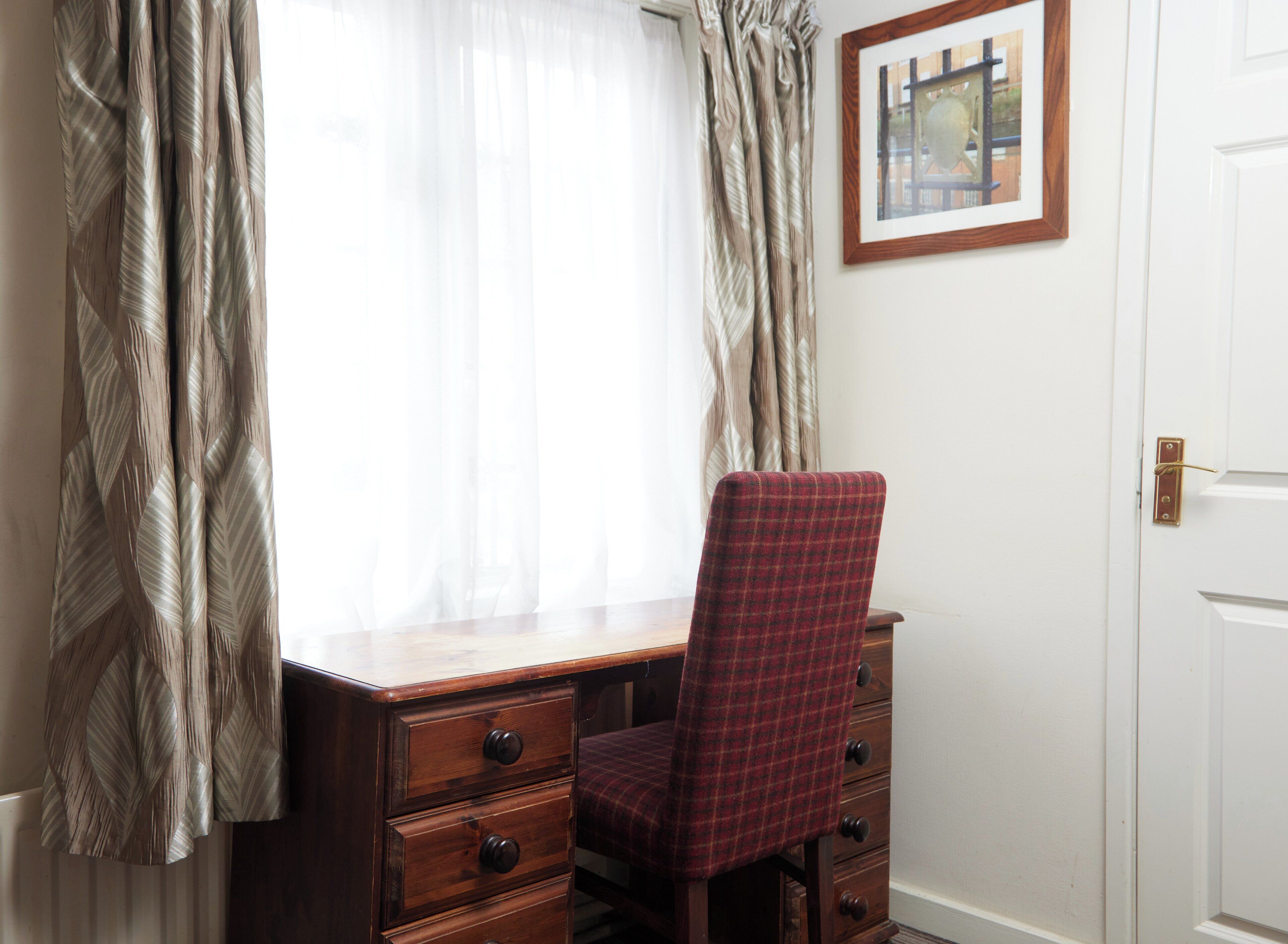 A room with a wooden desk, a plaid chair, and a framed picture on the wall.