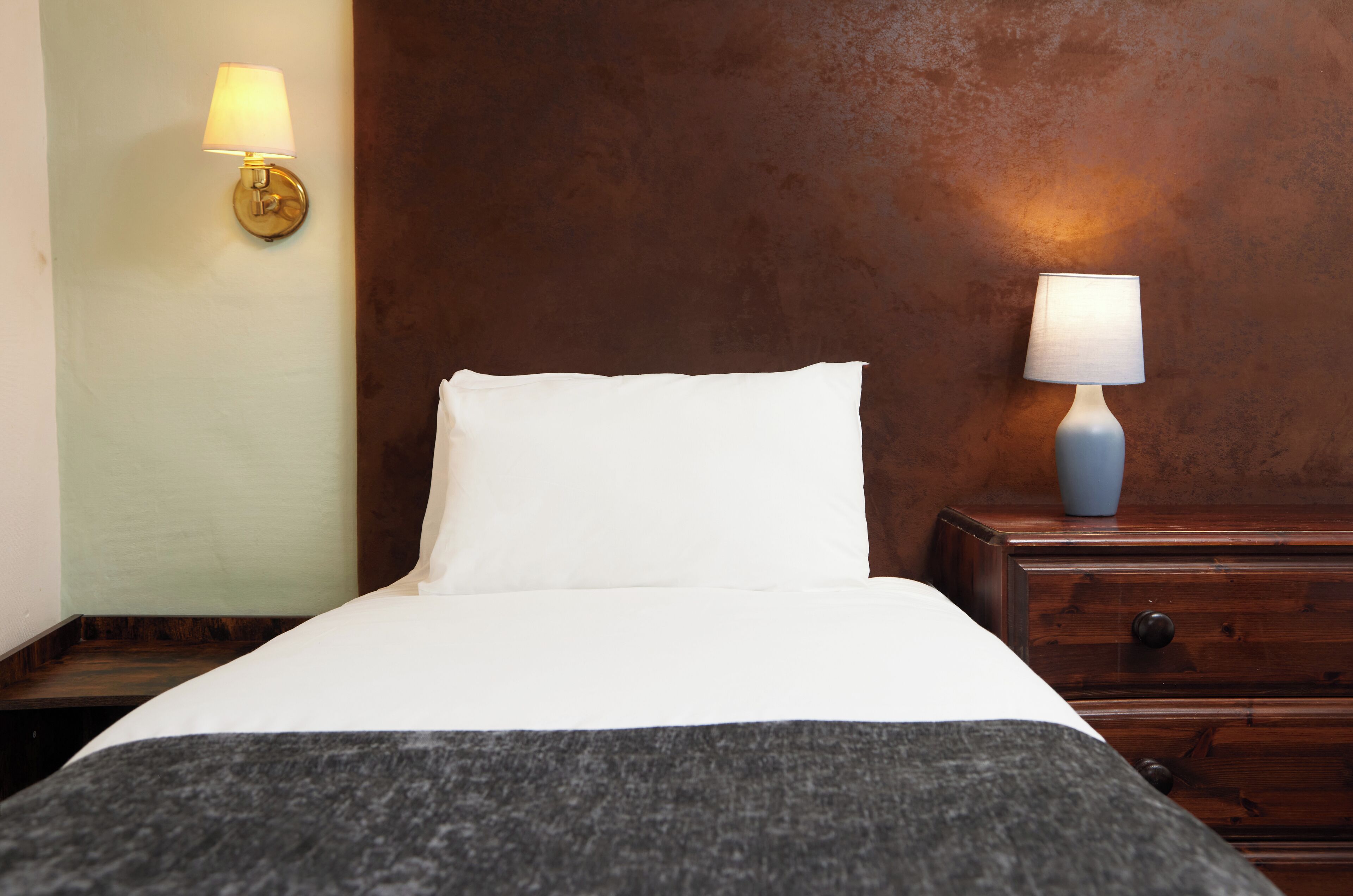 Una cama bien hecha con sábanas blancas y una manta gris, una mesita de noche de madera con una lámpara, y un aplique en la pared.