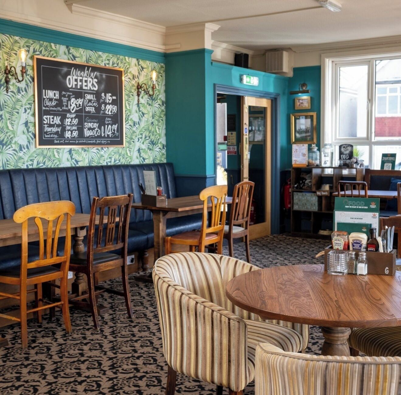A restaurant interior with a teal wall, a blackboard with weekly offers, wooden furniture, and a patterned carpet.