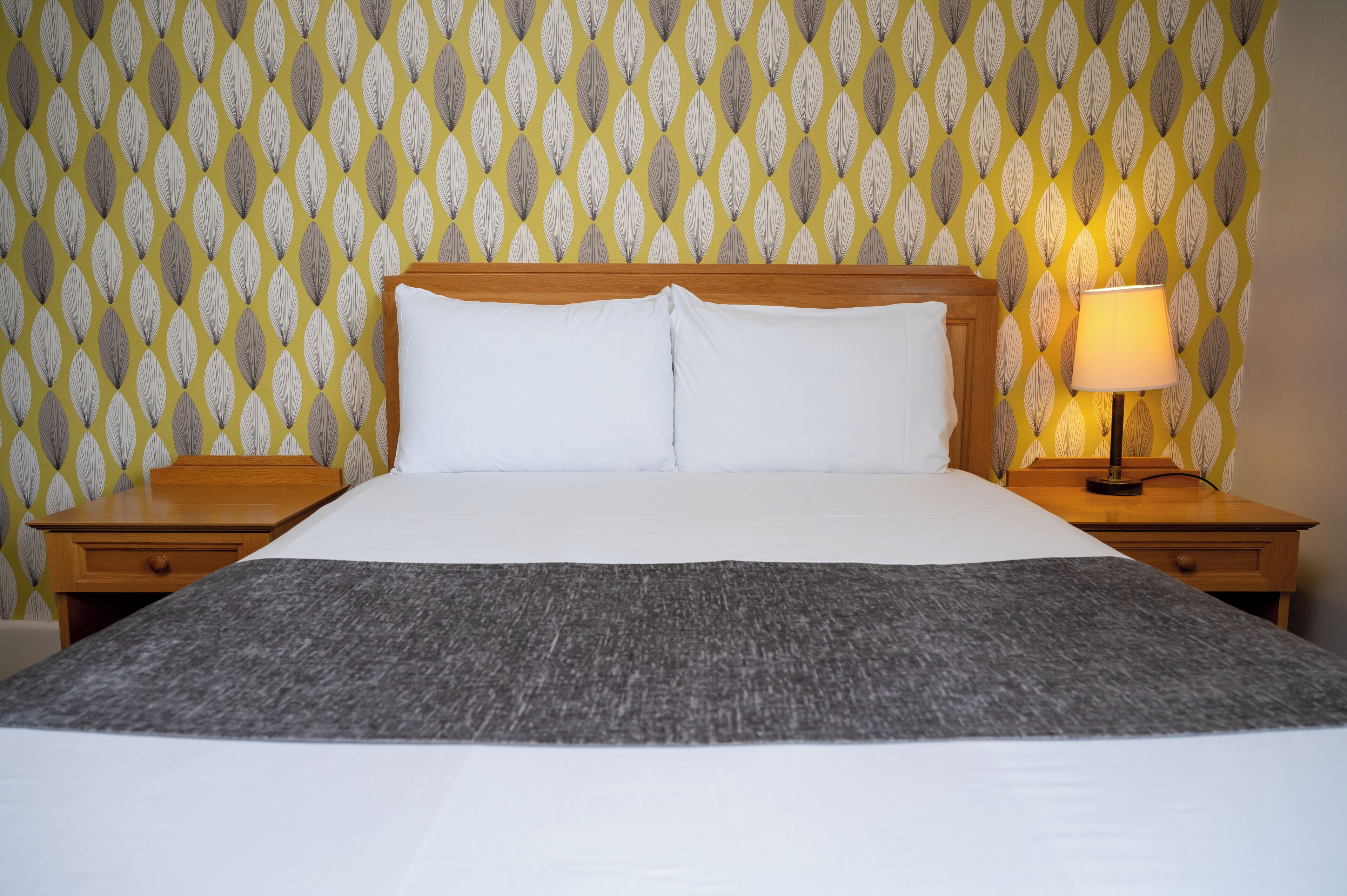 Una cama bien hecha con sábanas blancas y un plaid gris, flanqueada por mesitas de noche de madera con lámparas, sobre un fondo de papel tapiz estampado.
