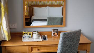 Quarto de hotel com escrivaninha de madeira, espelho, cama com roupa de cama branca, bandeja com uma caneca, controle remoto e um cartão sobre a escrivaninha.