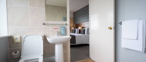 A modern bathroom with a toilet, sink, and mirror. A door leads to a bedroom with a bed and a patterned wall.