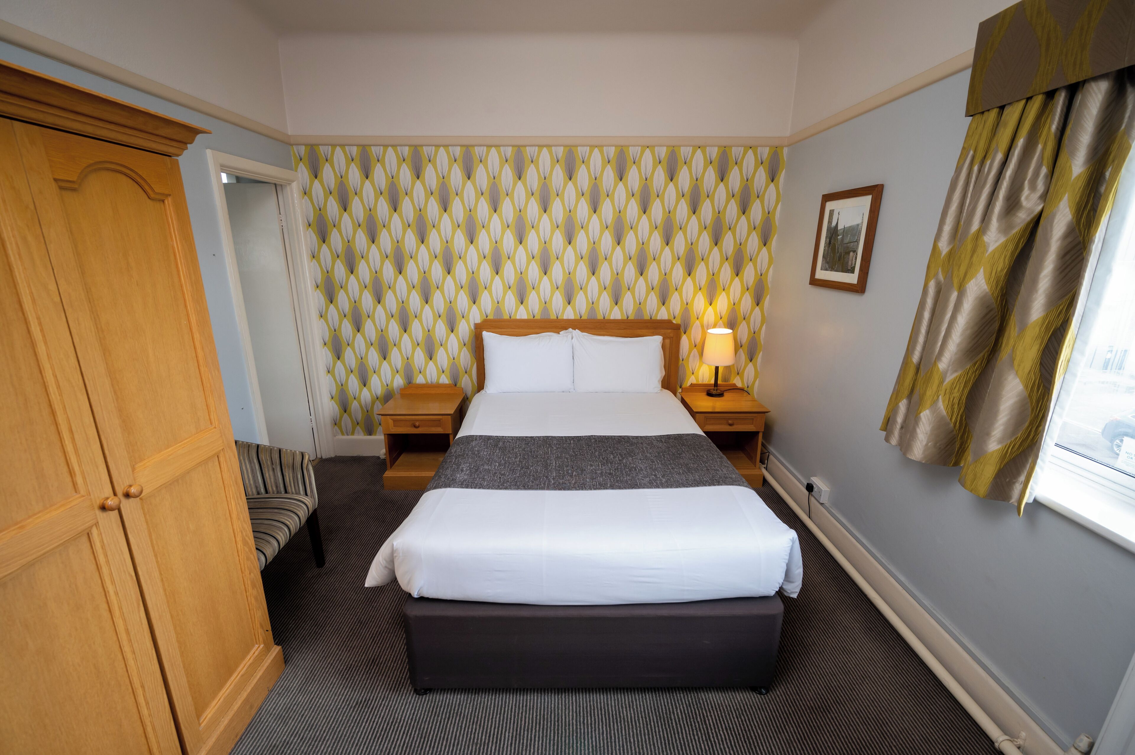 Une chambre d’hôtel avec un grand lit, une armoire en bois, un canapé à rayures, un papier peint à motifs et un tableau encadré au mur.