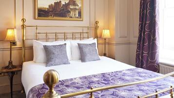 A neatly made bed with a purple patterned bedspread, two bedside lamps, and a painting above the bed.