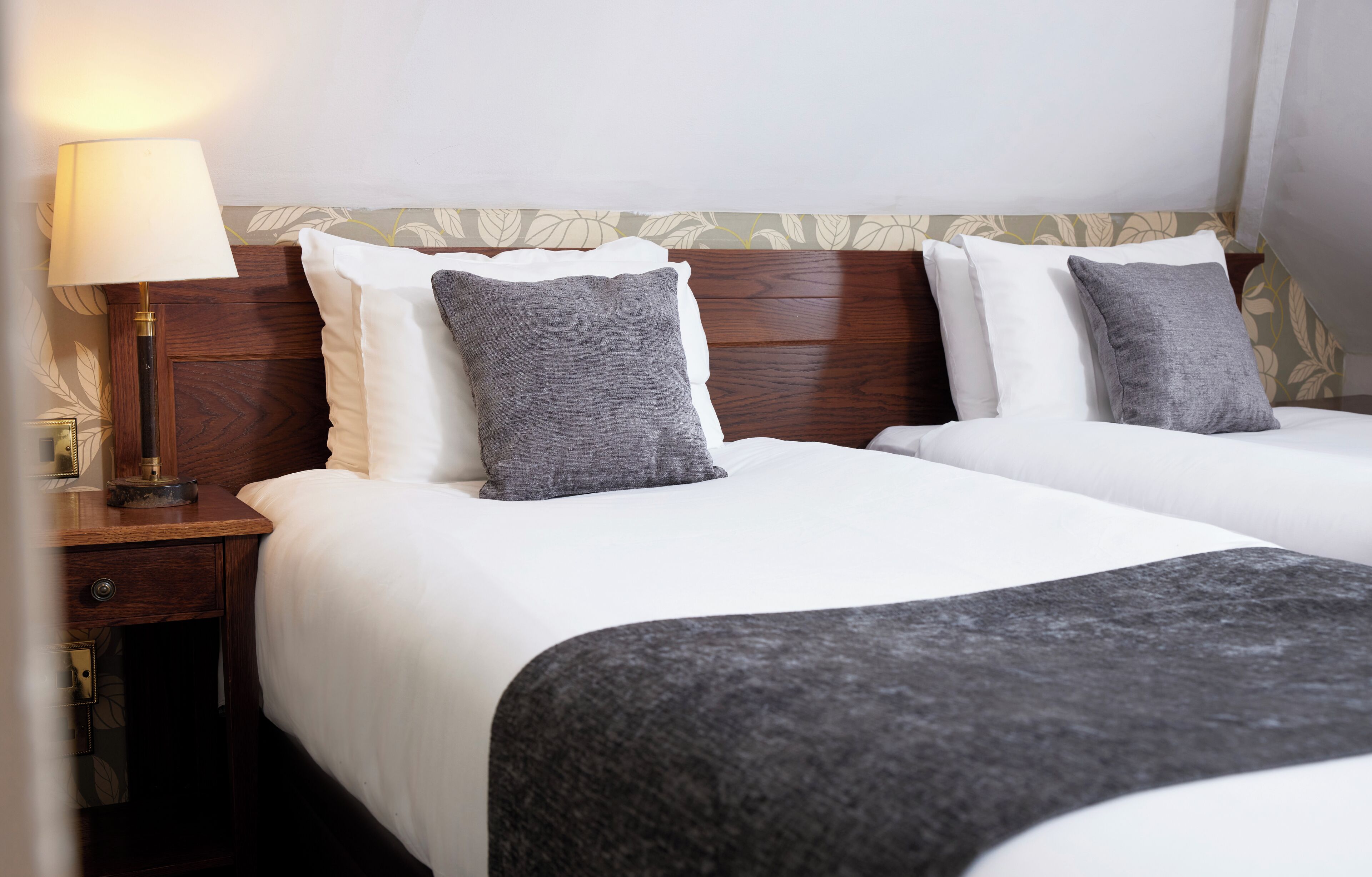 Ein Hotelzimmer mit zwei Betten, einem Nachttisch, einer Lampe und einem blumengemusterten Kopfteil.