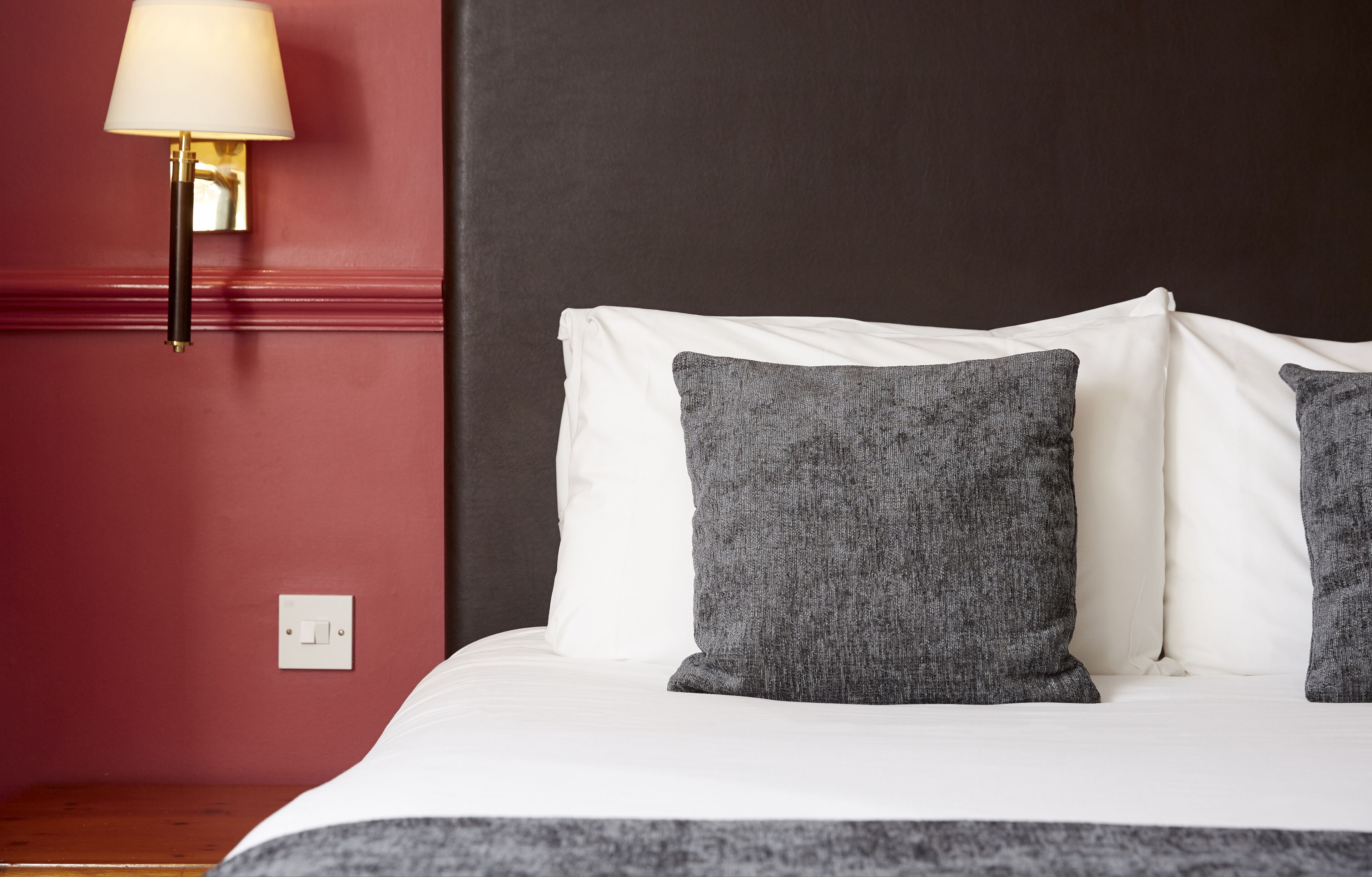 Una cama con almohadas blancas y grises, una lámpara fijada a la pared y un enchufe en una pared roja.