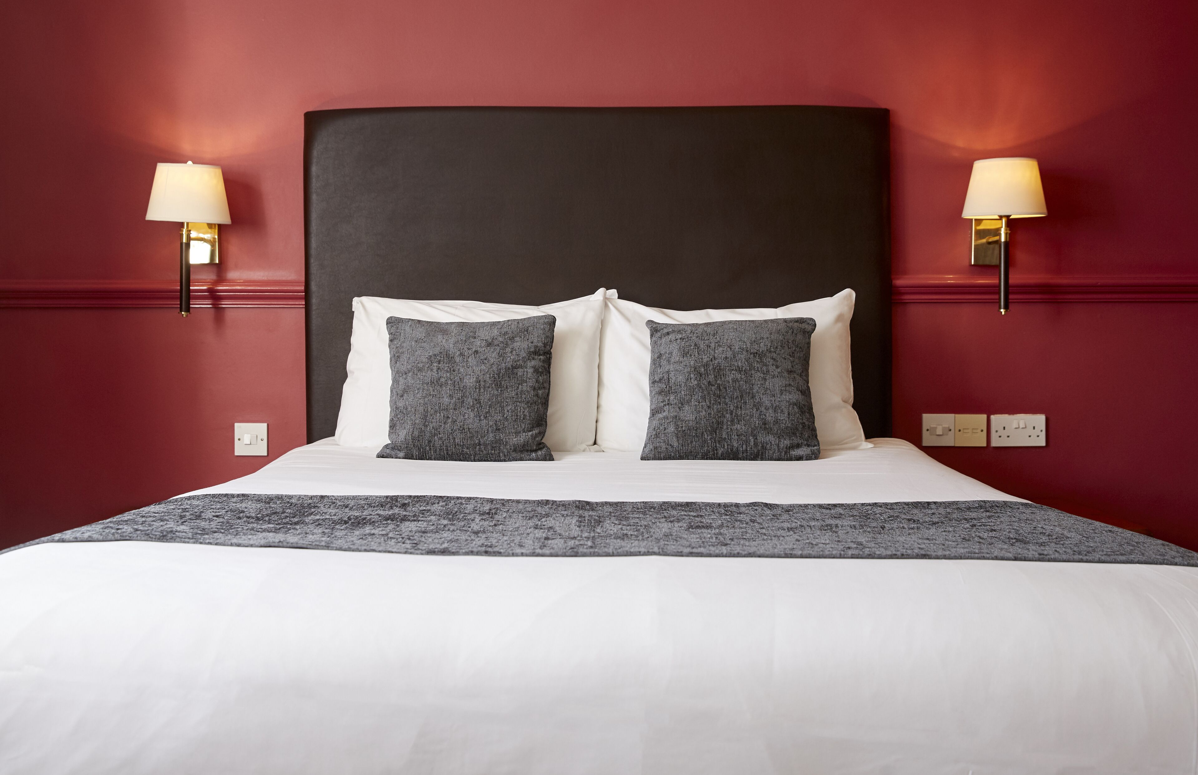 A neatly made bed with white and gray bedding, two wall-mounted lamps, and a red wall.