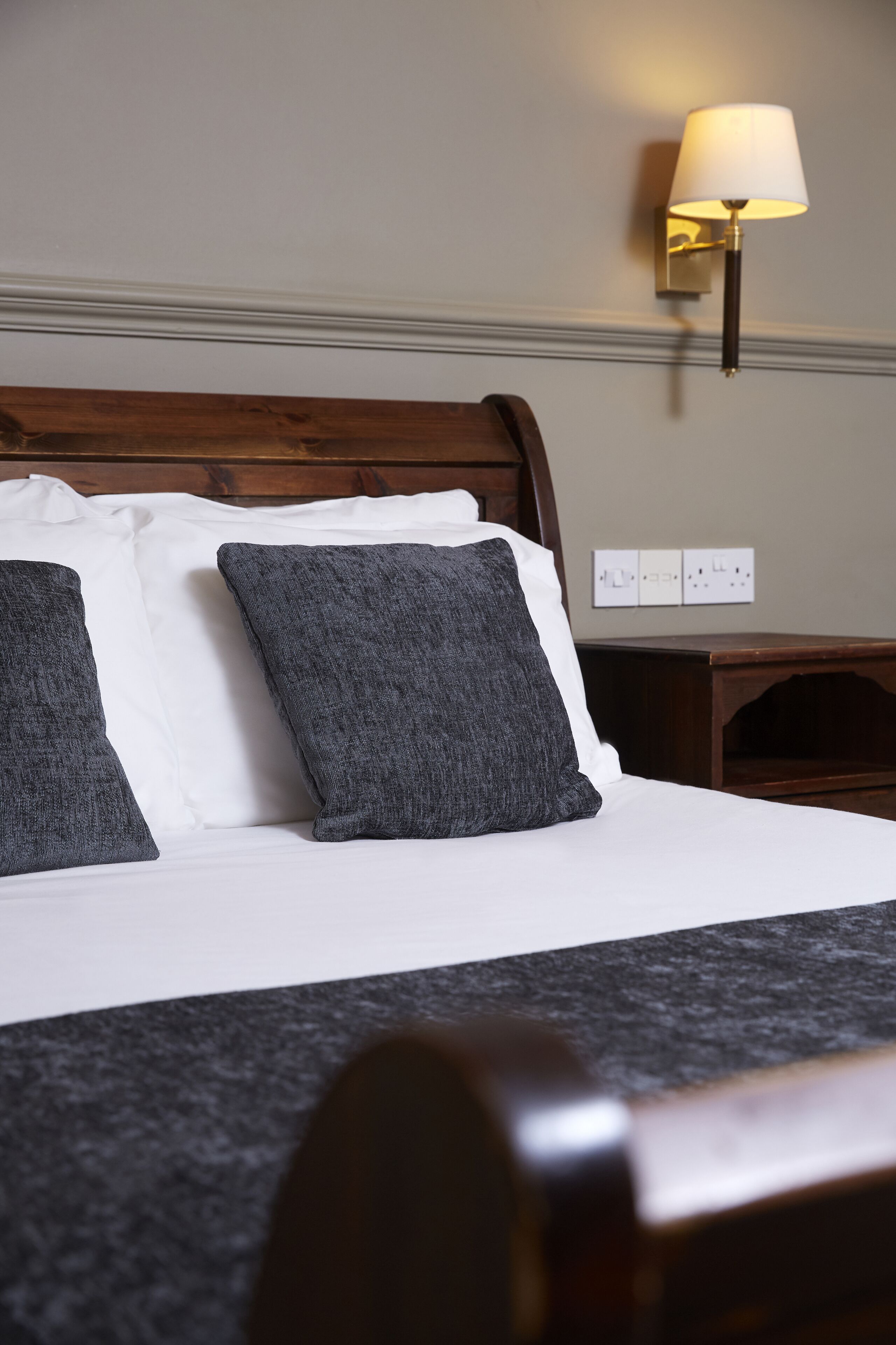 Una cama bien hecha con almohadas blancas y grises, un cabecero de madera y una lámpara fijada a la pared.