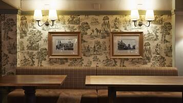 Interior de un restaurante con una zona de estar con bancos de madera, cuadros en la pared y dos lámparas de pared.