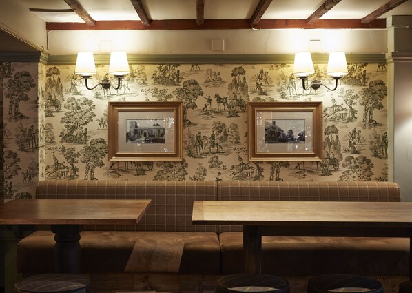 A restaurant interior with a wooden bench seating area, framed pictures on the wall, and two wall-mounted lamps. - Dog House  by Chef and Brewer Collection (Abingdon)
