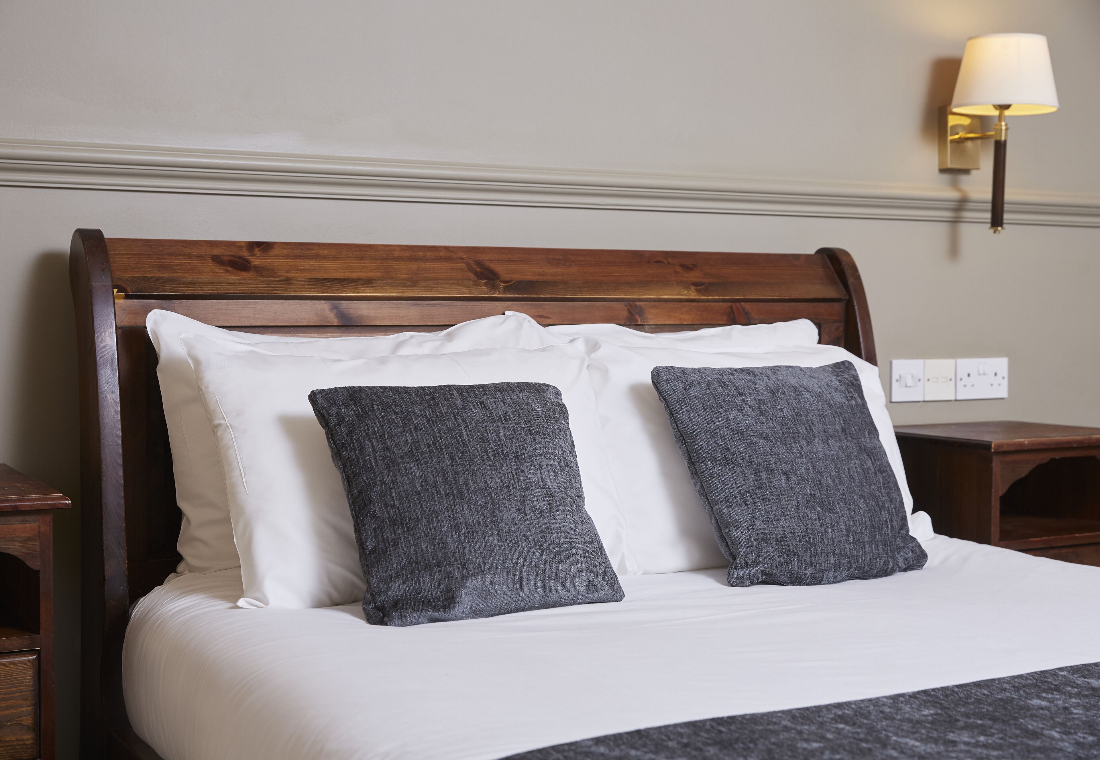 Un lit bien fait, avec des oreillers blancs et gris, une tête de lit en bois et une lampe fixée au mur.