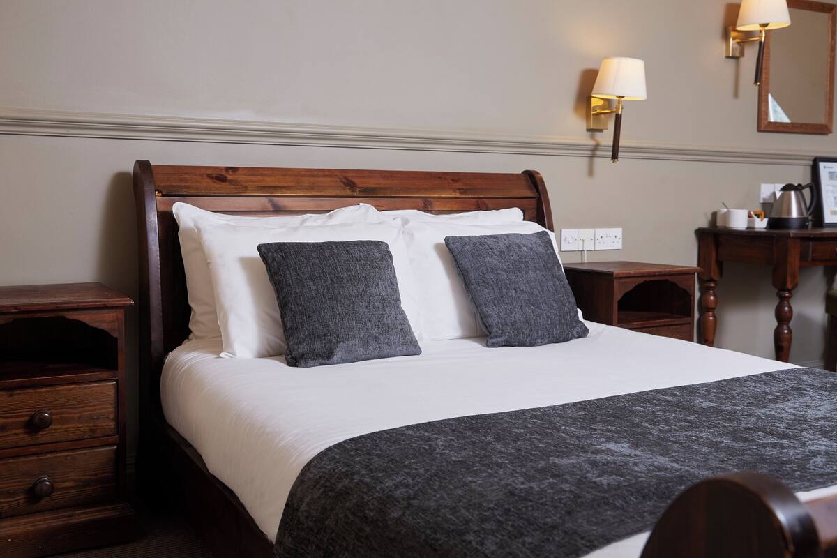 A neatly made bed with white and grey bedding, two bedside tables, a nightstand, and wall-mounted lamps.