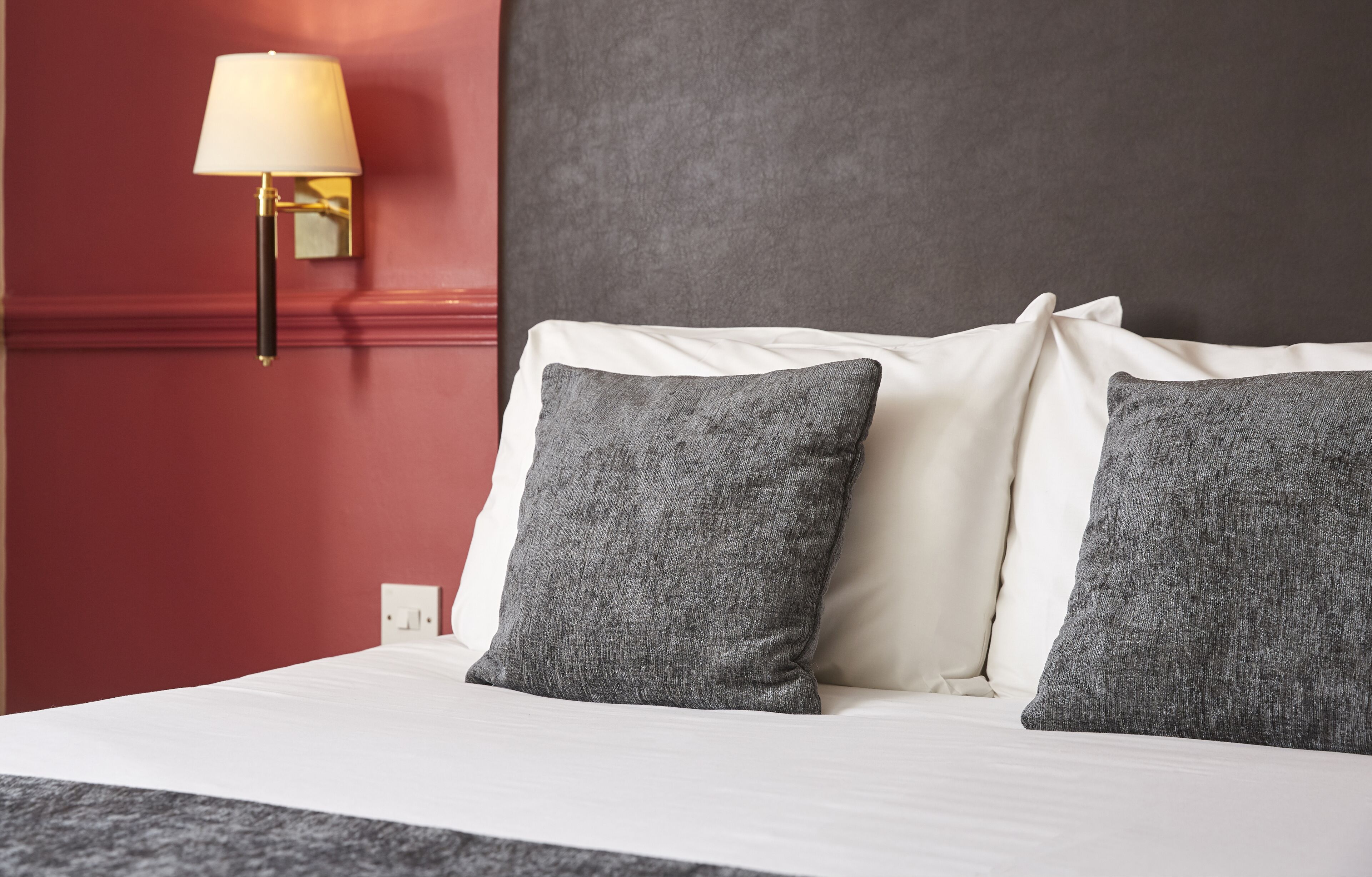 A hotel room with a bed, two pillows, a wall-mounted lamp, and a red accent wall.