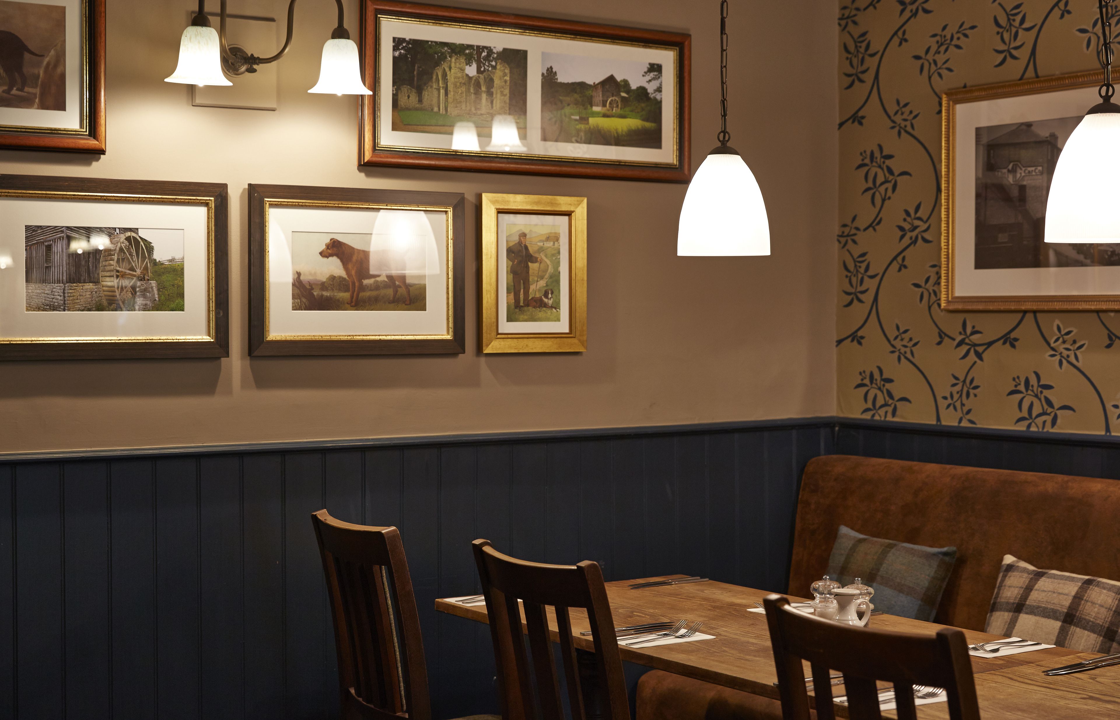 A dining area with a wooden table, chairs, and a bench. There are framed pictures on the wall and pendant lights above the table.
