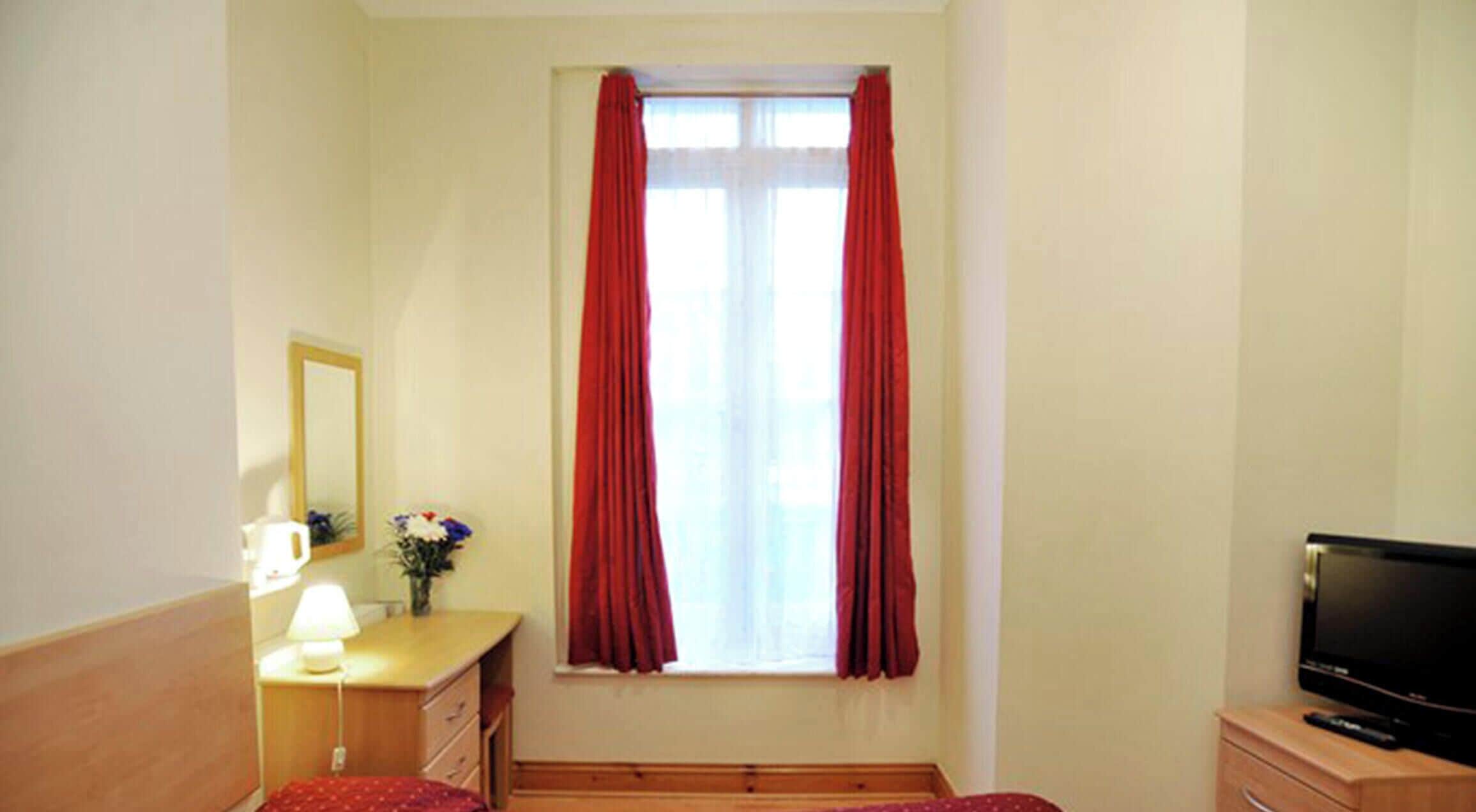 Une chambre à coucher avec un lit, un bureau avec une lampe, une télévision et une fenêtre avec des rideaux.