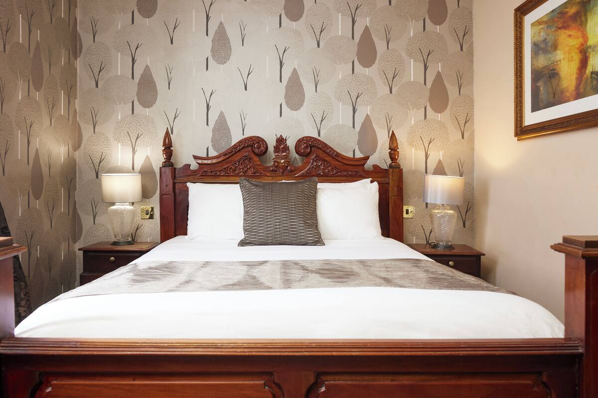 A neatly made bed with a wooden headboard, two bedside tables with lamps, and a patterned wallpaper with tree designs.