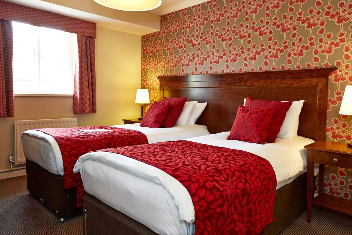 A hotel room with two beds, a wooden headboard, a floral wallpaper, a window with curtains, and a nightstand with a lamp.