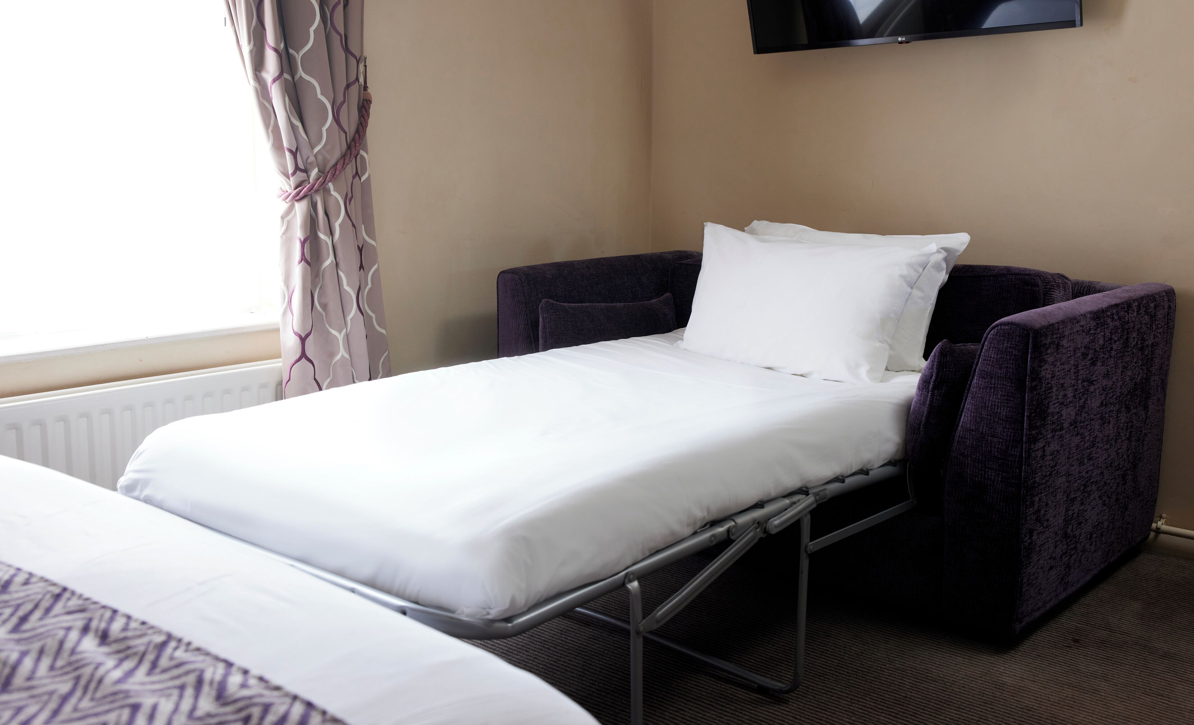 Una cama individual con estructura metálica, un sillón morado y una almohada blanca.