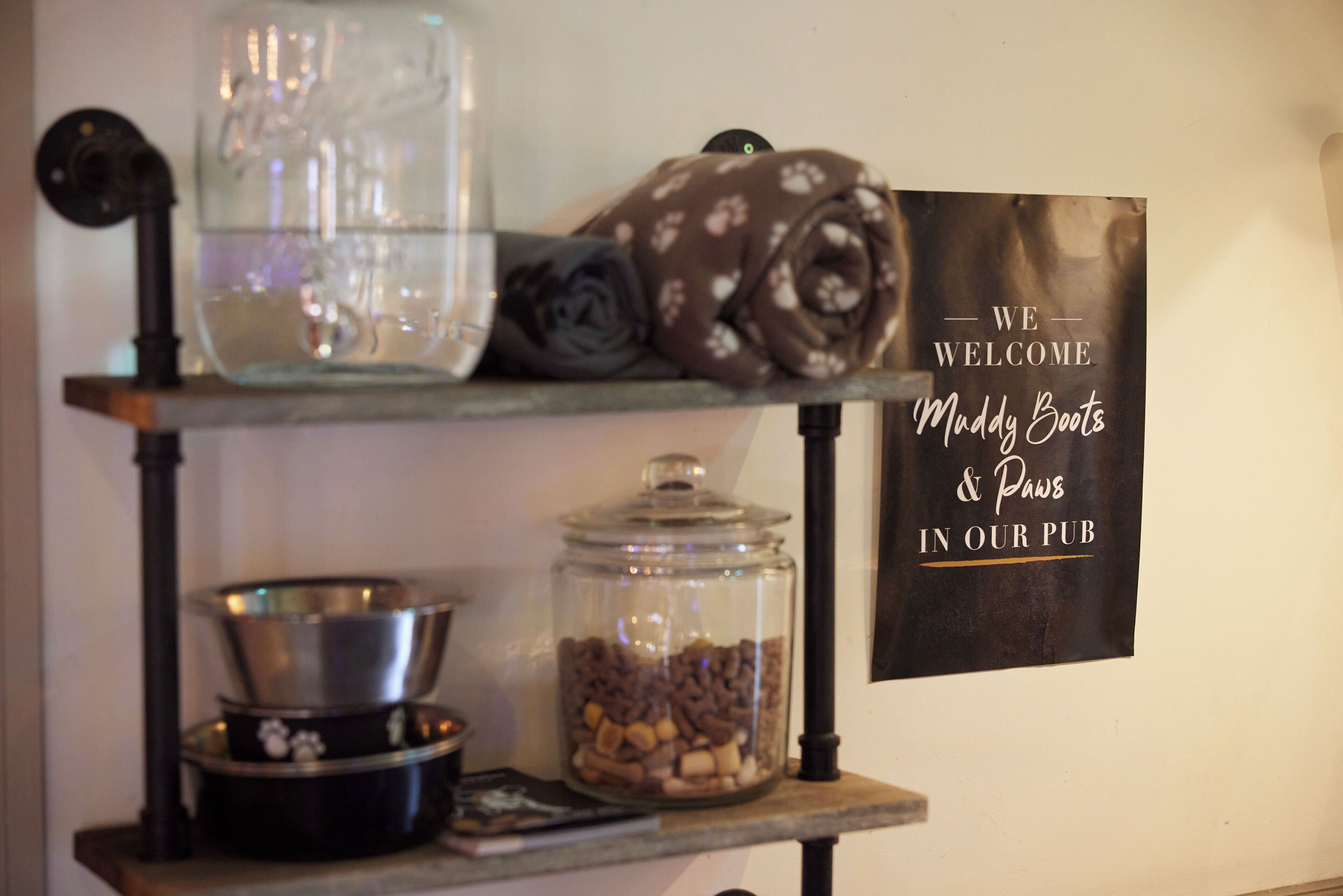 A shelf with a glass jar of dog treats, a rolled-up blanket, and a metal bowl. A sign reads "We welcome Muddy Boots & Paws in our pub."