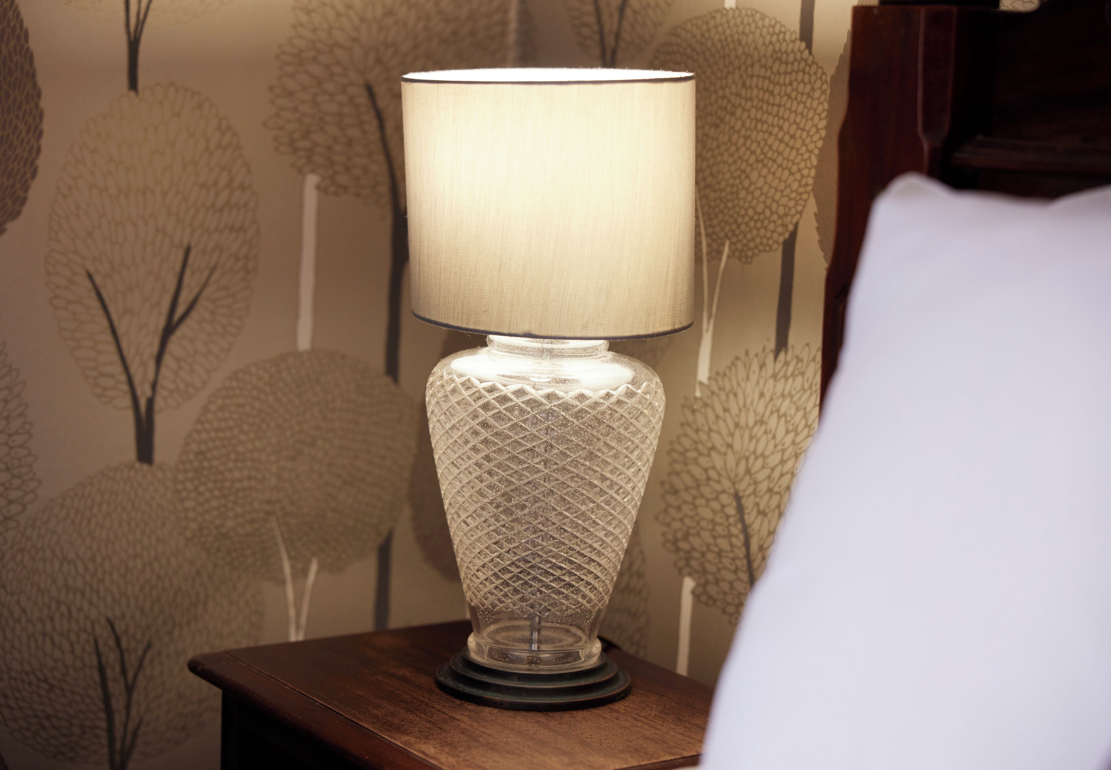 A table lamp with a textured base and a cylindrical shade, placed on a wooden table against a wall with a tree pattern wallpaper.