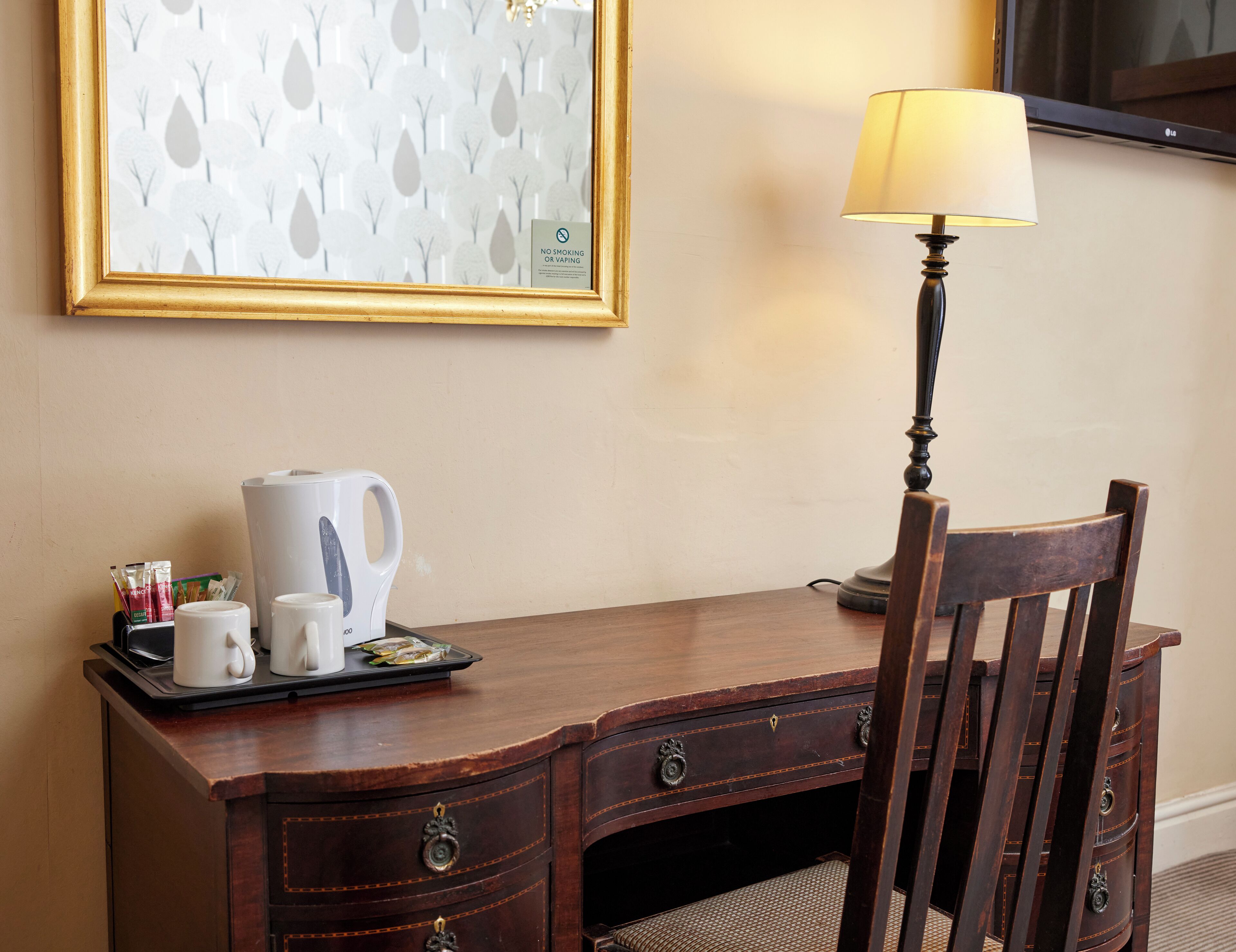A wooden desk with a lamp, a framed picture, and a tray with a kettle and cups.