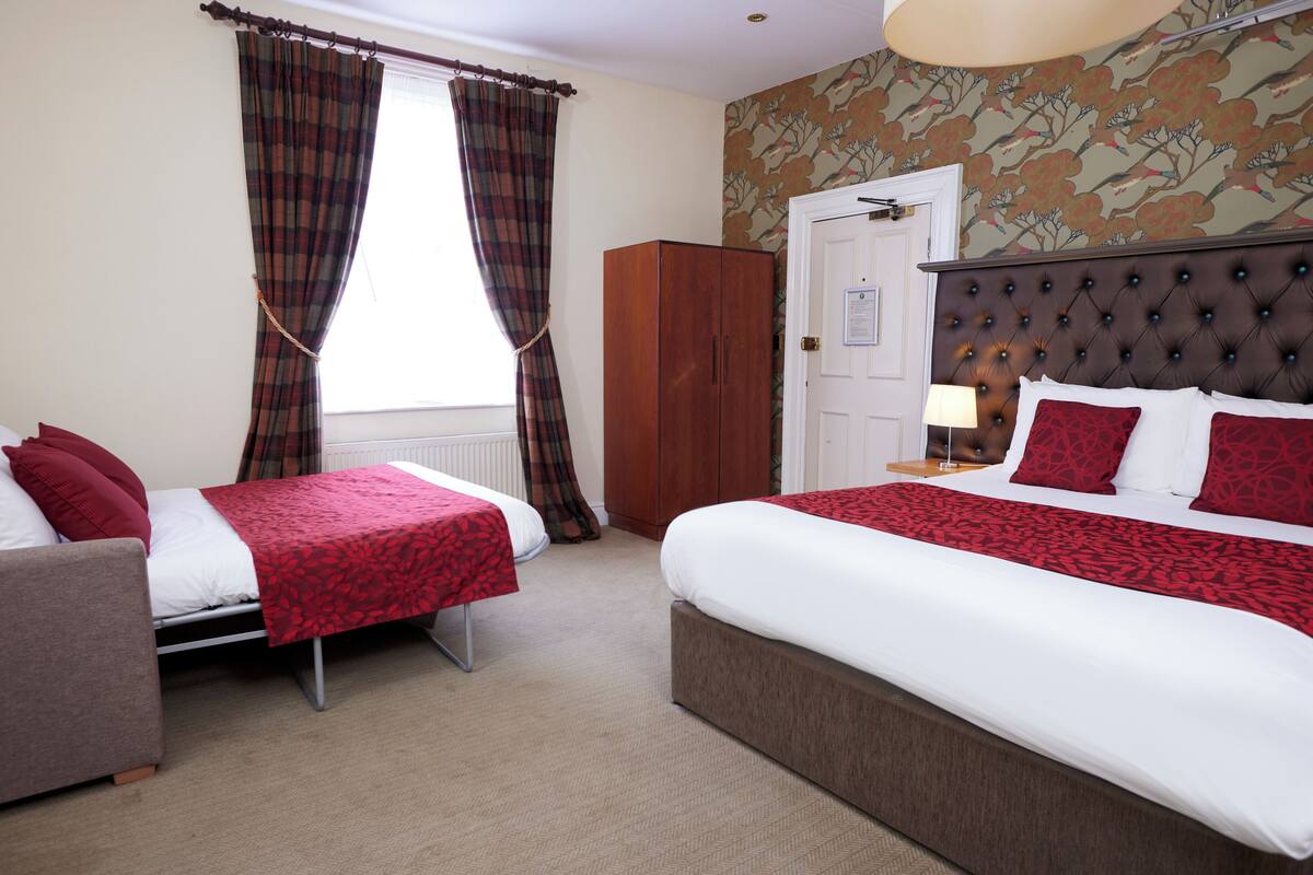 A hotel room with two beds, a wardrobe, a desk with a lamp, and a window with curtains.