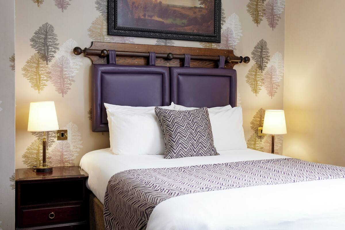 A neatly made bed with a patterned bedspread, flanked by two bedside tables with lamps, set against a wall with leaf-pattern wallpaper and a framed picture above the bed.