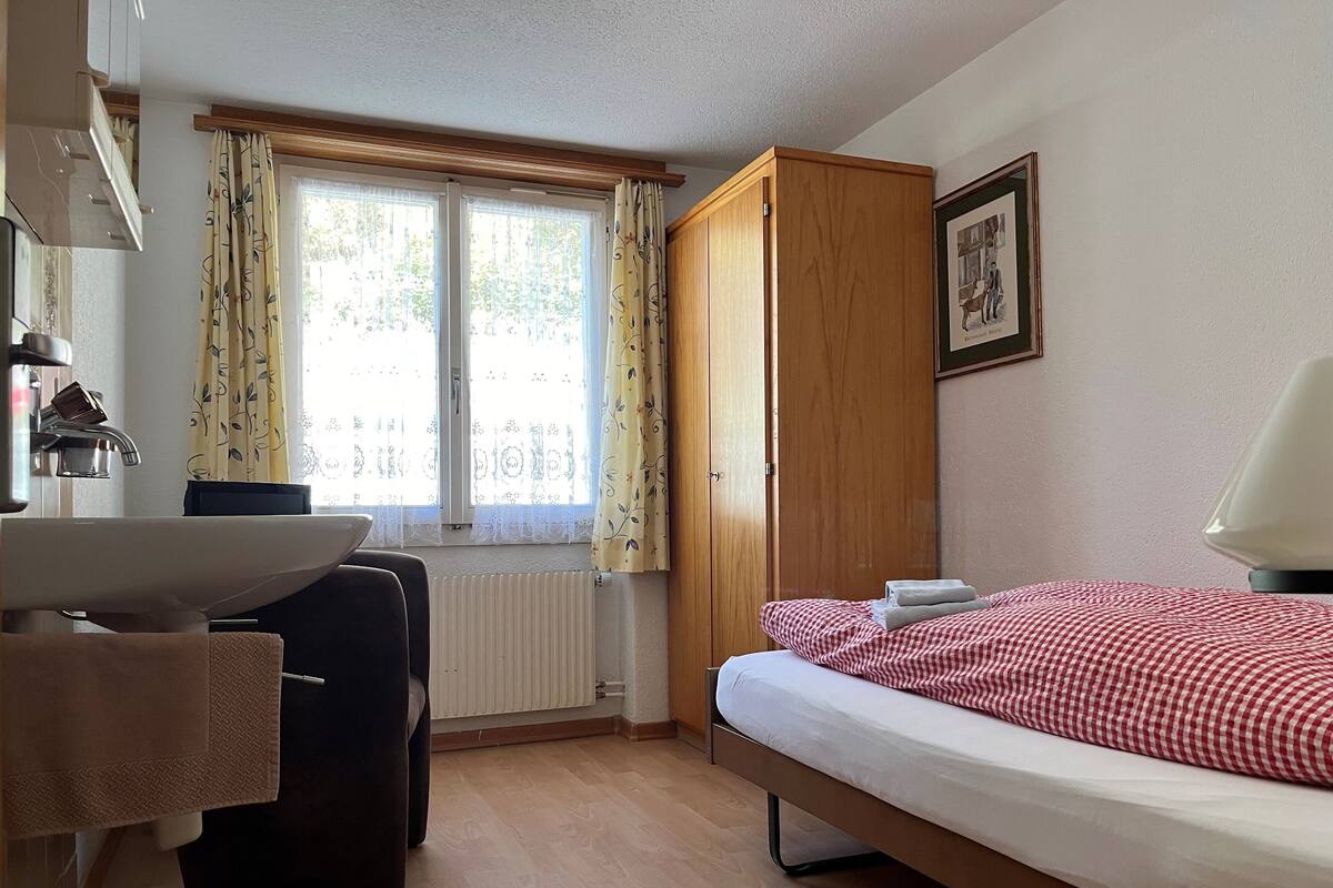 Ein Schlafzimmer mit Bett, Holzschrank, Heizkörper, Fenster mit Vorhängen, Waschbecken und Spiegel.