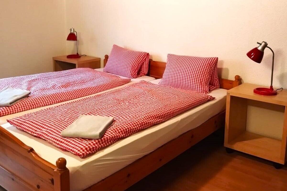 Ein hölzernes Bett mit karierter Bettwäsche, zwei Nachttische und je eine rote Lampe auf beiden Seiten.